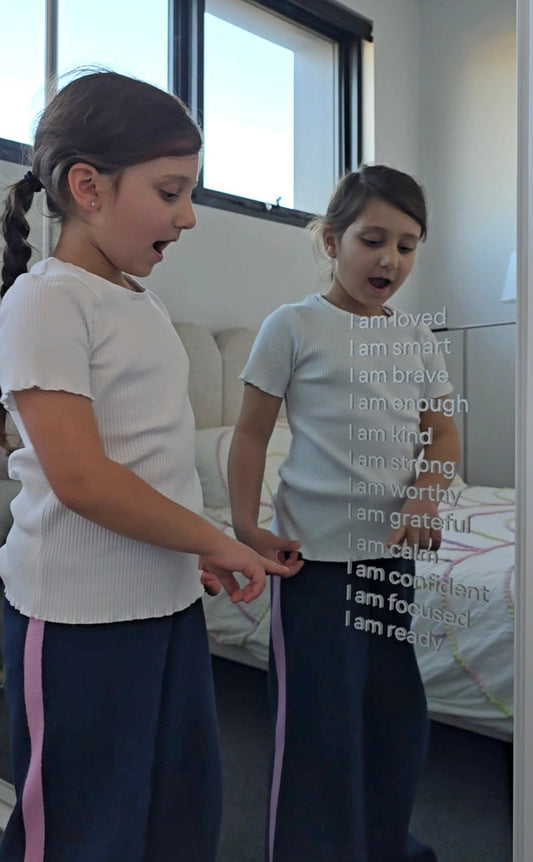 one young girls standing in a bedroom, looking at a mirror. the girl wears a light gray t-shirt and dark navy pants with white text down the front reading 'I am loved.