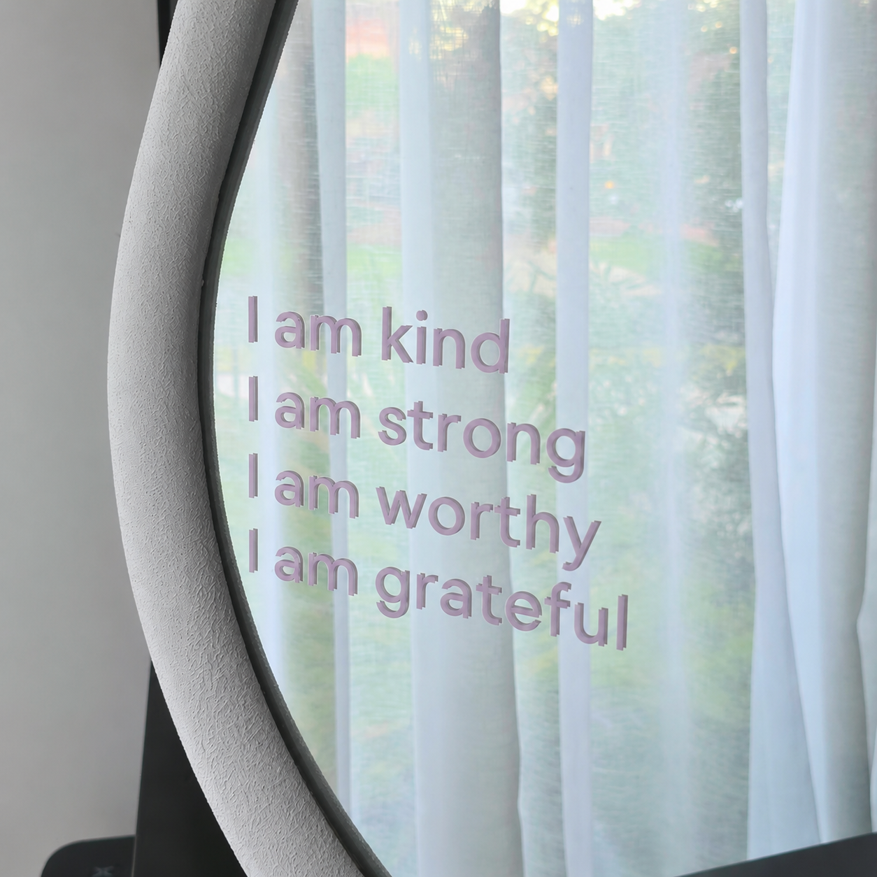 A close-up of a curved glass surface with the phrase 'I am kind I am strong I am worthy I am grateful' printed in a light lavender-purple font, set against sheer white curtains and a blurred outdoor background.