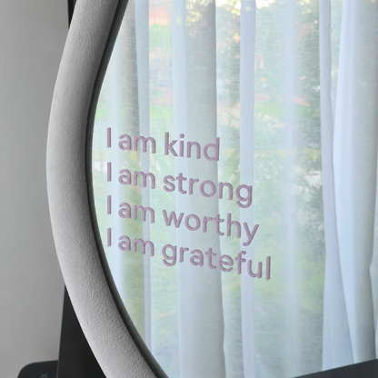 A close-up of a curved glass surface with the phrase 'I am kind I am strong I am worthy I am grateful' printed in a light lavender-purple font, set against sheer white curtains and a blurred outdoor background.