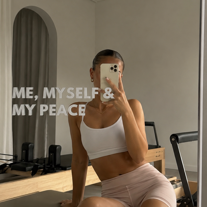 A woman takes a mirror selfie in a minimalist studio, wearing a white sports bra and light pink athletic shorts. She is sitting on a Pilates reformer machine.